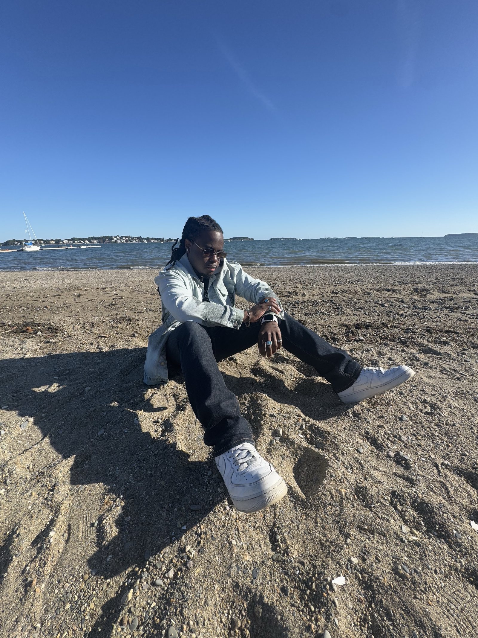 Marshall Delpé sitting on a sandy beach in a denim jacket with the sea and a sailboat behind him
