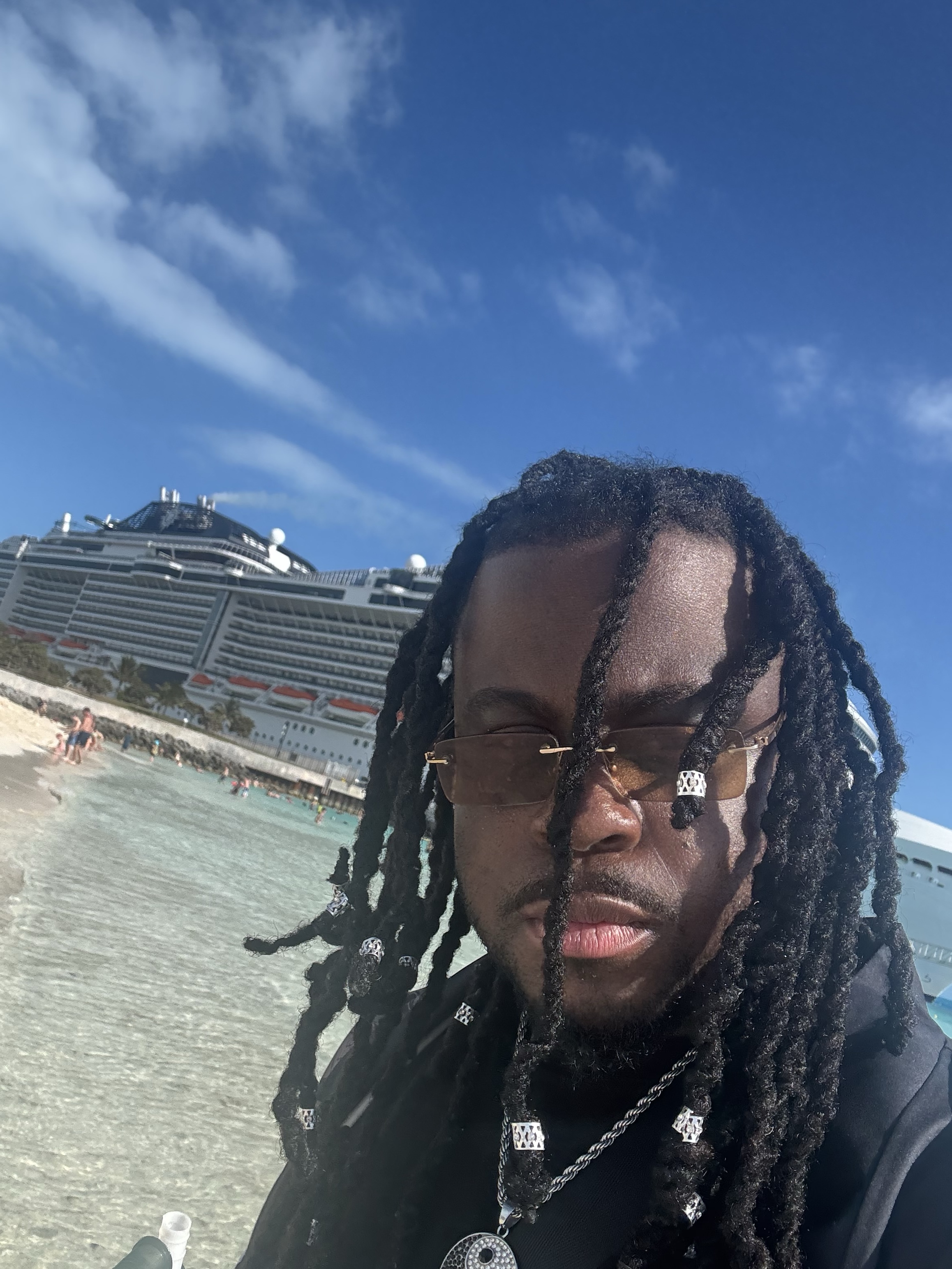 Marshall Delpé taking a selfie by the shore with a large cruise ship and blue sky behind him